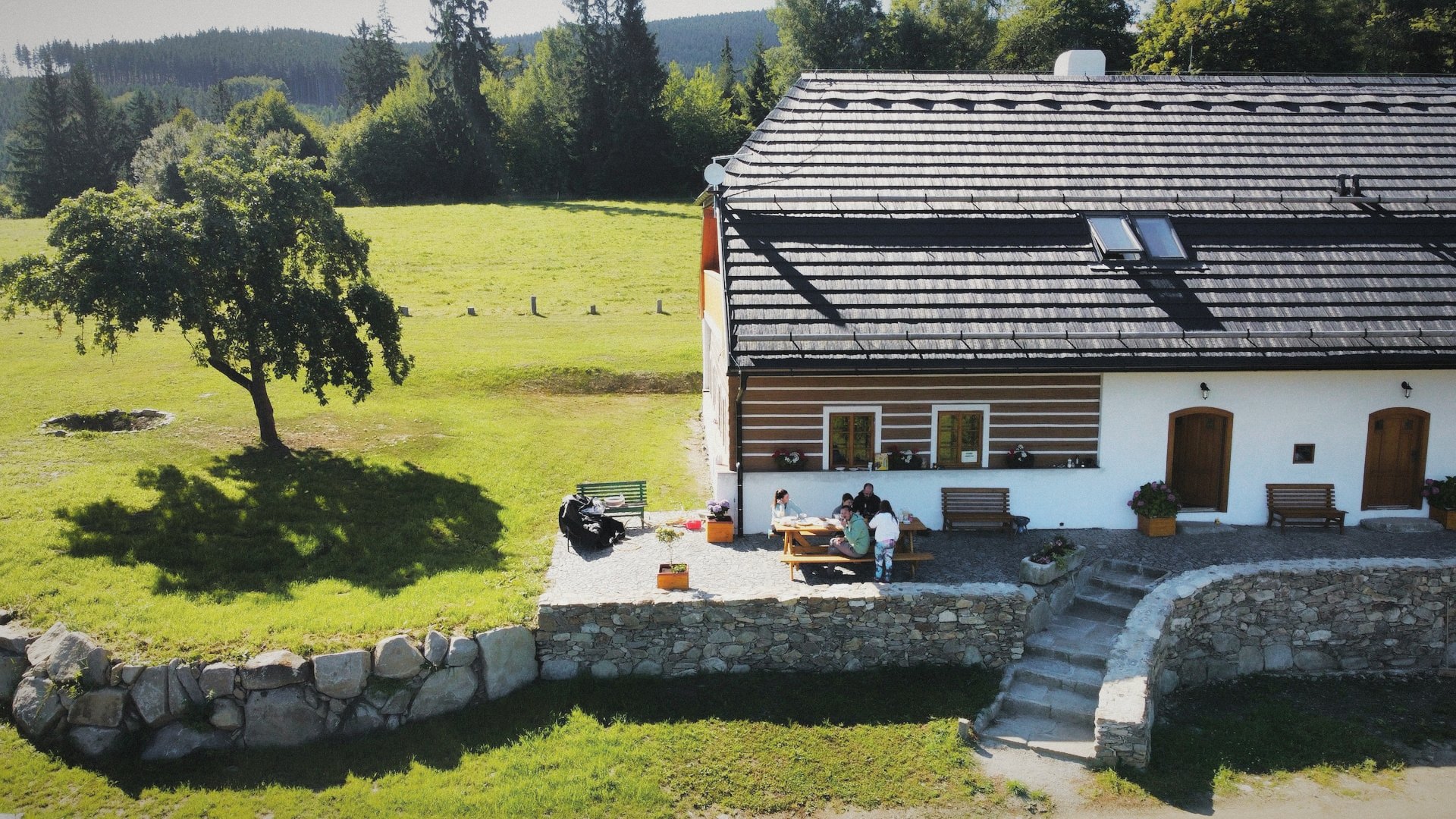 Roubená chalupa Kukanda s bílými spárami a šindelovou střechou na louce, před ní kamenná terasa a jabloň