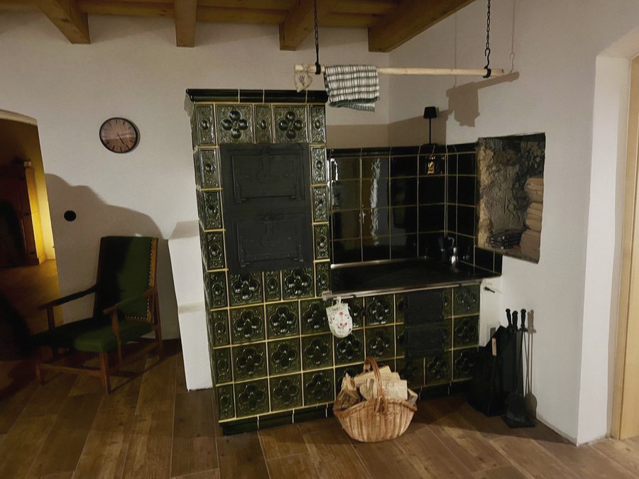 Zelená kachlová kamna s plotnou, vedle křeslo a koš na dřevo